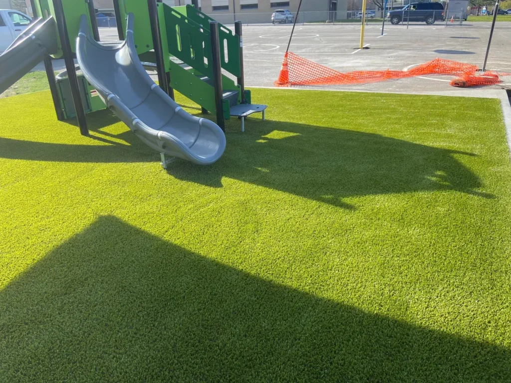 Newly installed bright green artificial playground turf surrounds a green and gray slide structure at a Fort Wayne school.