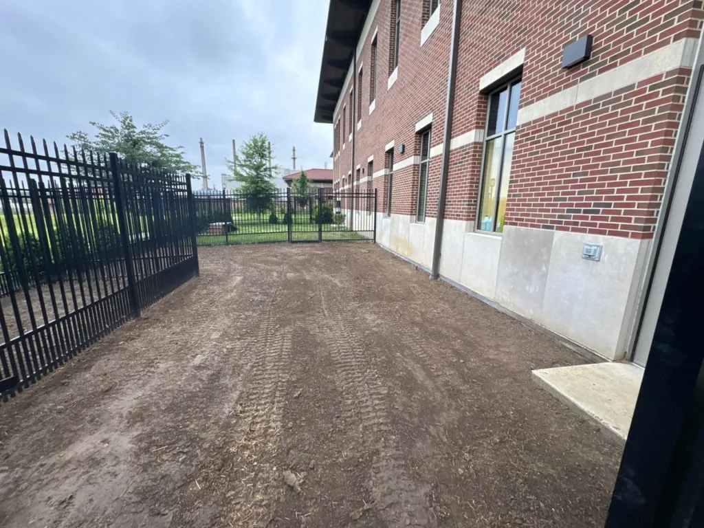 Freshly graded bare soil area beside brick veterinary clinic building enclosed by black iron fence, prepped for K9Grass installation.