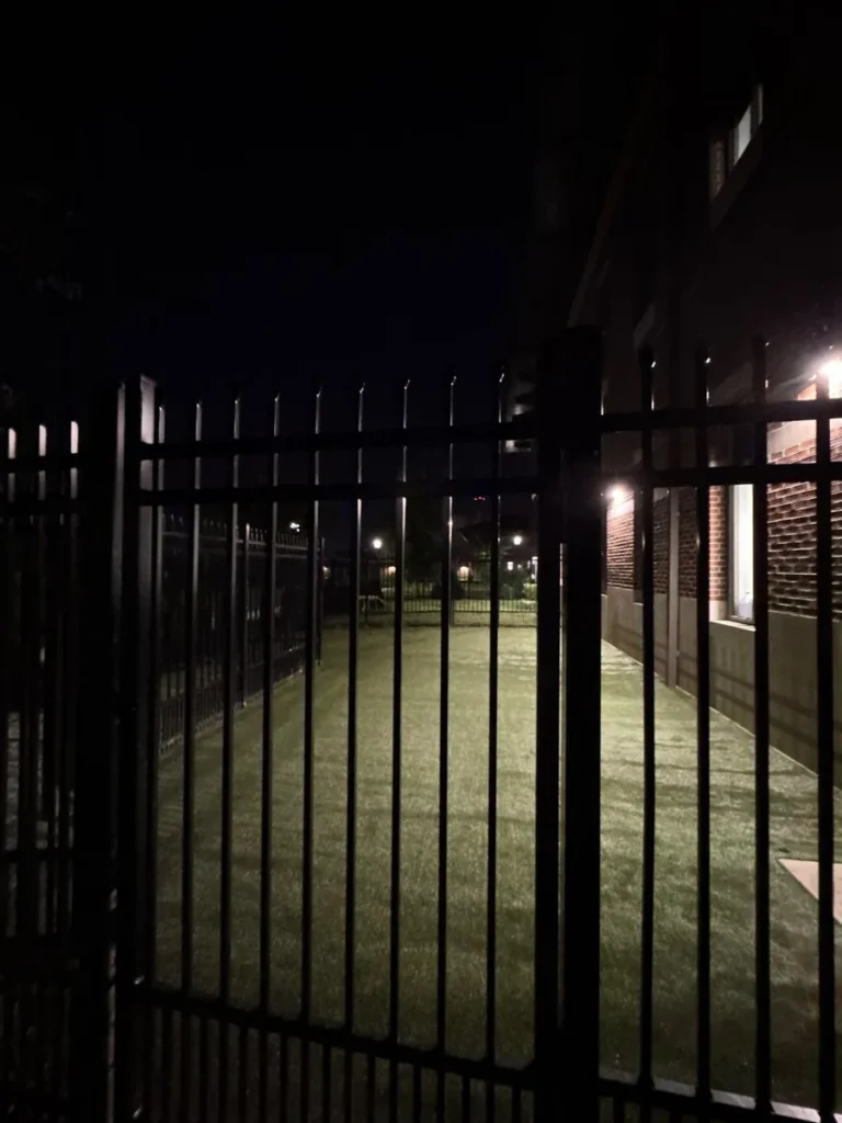 Night view of K9Grass Elite artificial turf dog run enclosed by black iron security fencing beside brick veterinary clinic building.