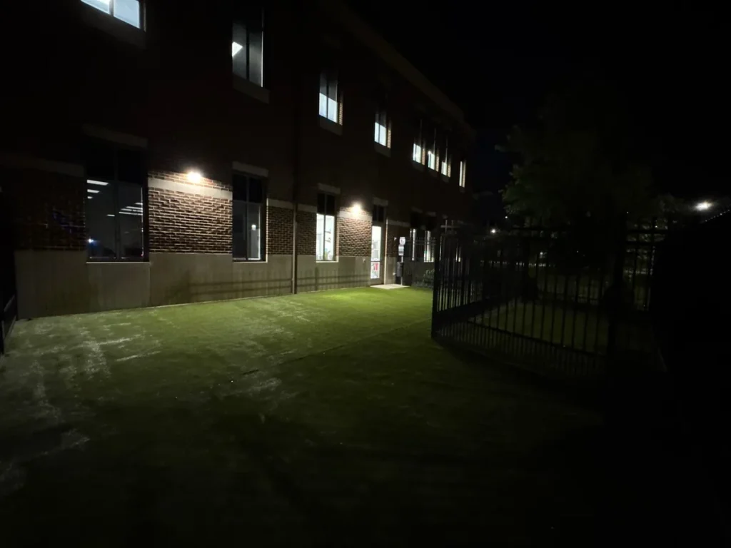 Nighttime K9Grass Elite artificial turf dog run at veterinary clinic, enclosed by black iron fence beside brick building.
