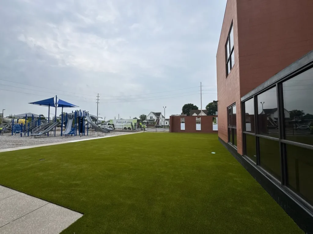Artificial turf playground area beside brick school building with blue shade structure and play equipment in background.
