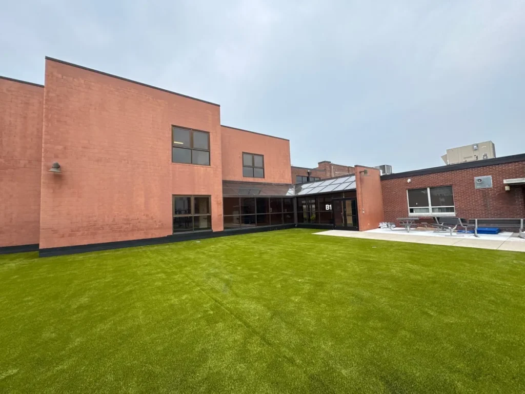 Artificial turf courtyard installed between brick school buildings with glass-covered walkway and adjacent patio seating area.