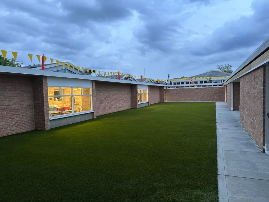 Artificial turf playground courtyard at Columbus elementary school, surrounded by brick buildings with yellow bunting flags overhead.