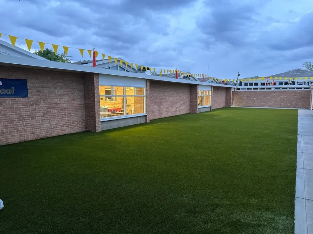 Playground Grass Ultra artificial turf installed at Columbus elementary school courtyard with brick building, large windows, and yellow bunting overhead.