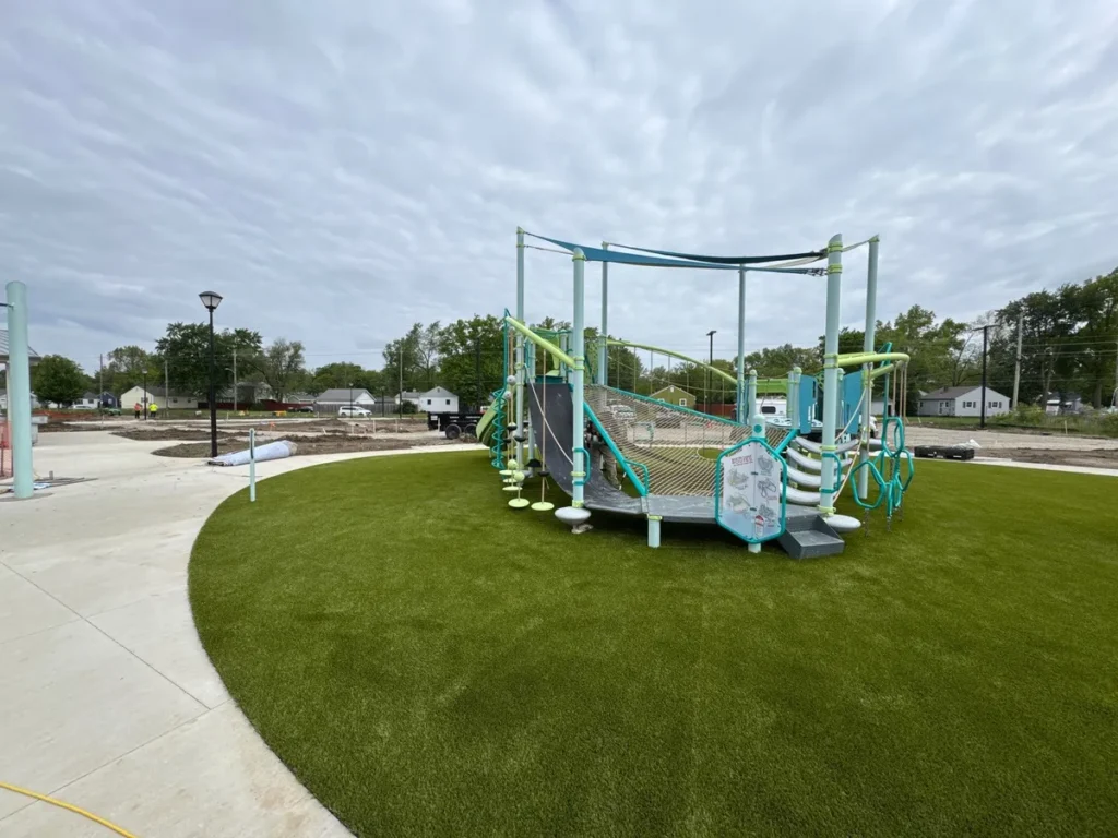 Playground Grass Ultra artificial turf surrounds modern teal playground structure at Fort Wayne school, under cloudy sky.