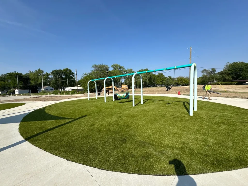 Circular artificial turf playground with teal swing set being installed at Fort Wayne school, construction equipment visible in background.