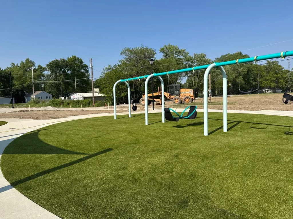 Newly installed Playground Grass Ultra artificial turf beneath teal swing set at Fort Wayne school playground, construction equipment visible in background.