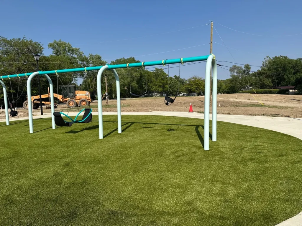 New playground swing set installed on Playground Grass Ultra artificial turf at Fort Wayne school, construction equipment visible in background.