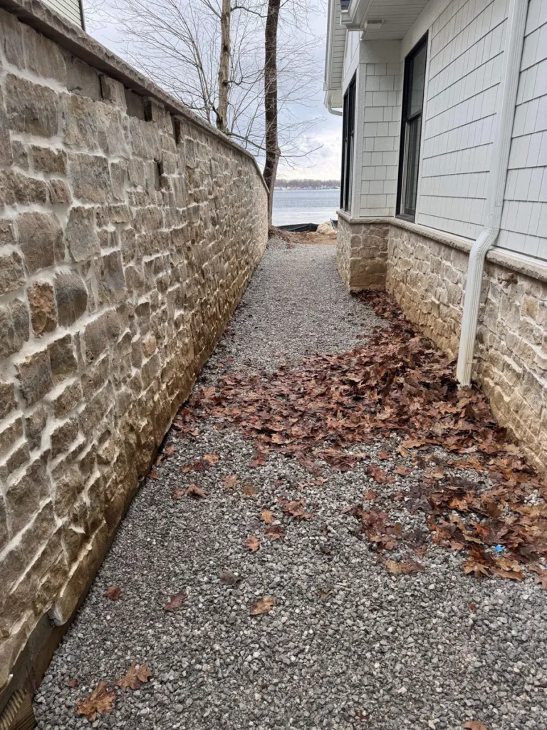 Narrow gravel pathway between stone retaining wall and white shingle house exterior, scattered fall leaves, waterfront view ahead.