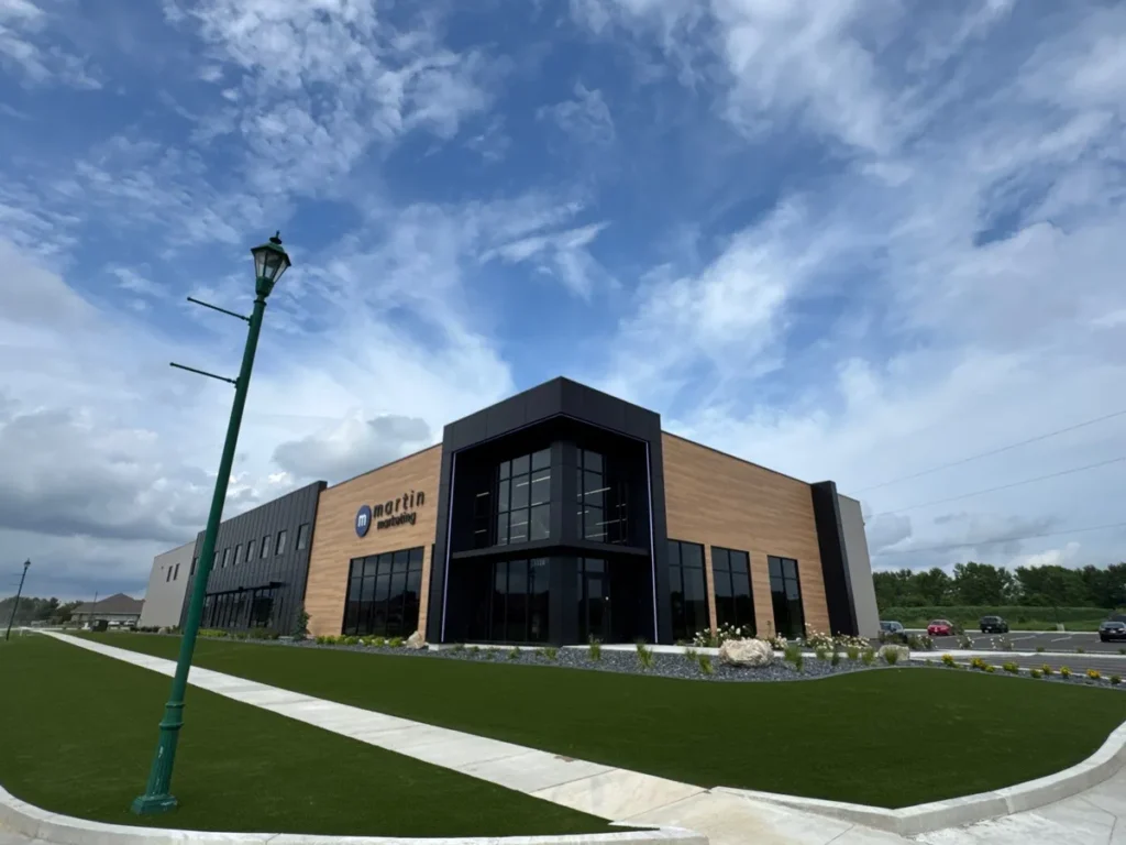 Lush artificial turf lawn with concrete pathways fronting modern two-story Martin Marketing commercial office building under blue sky.