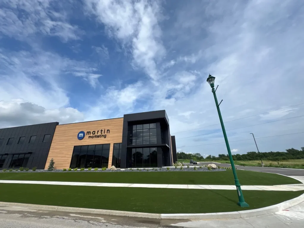 Lush artificial turf lawn fronting modern Martin Marketing office building with wood and dark metal facade under blue sky.