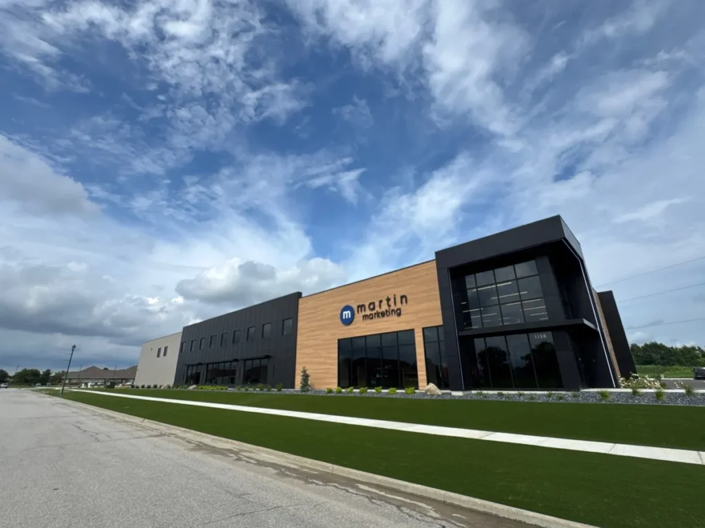 Lush artificial turf lawn fronting modern Martin Marketing commercial building with wood and dark metal facade under cloudy sky.