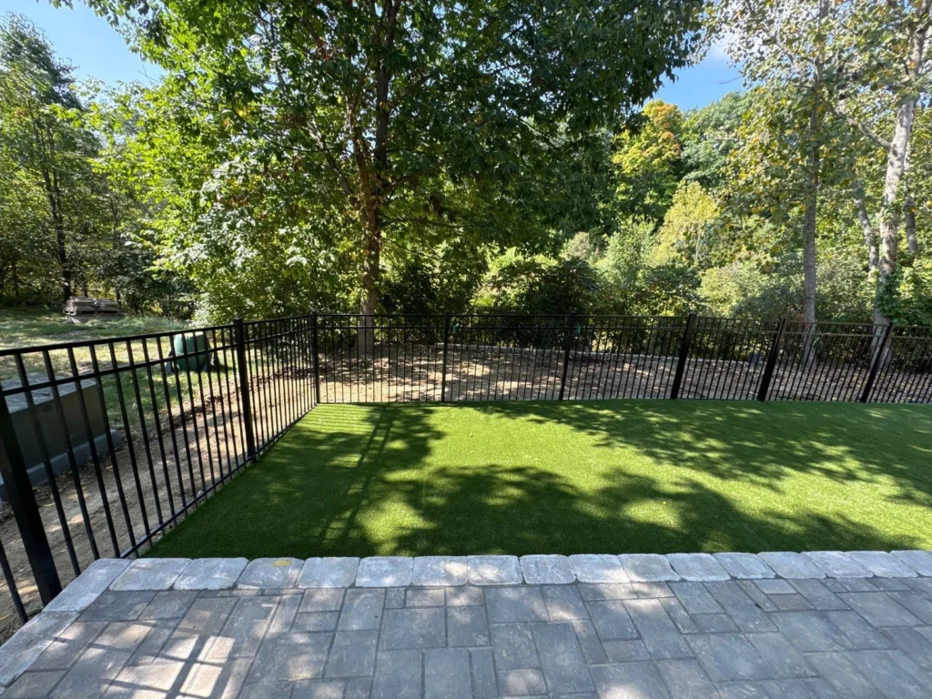 K9Grass Elite artificial turf pet yard with black aluminum fence, stone paver border, and wooded backdrop.