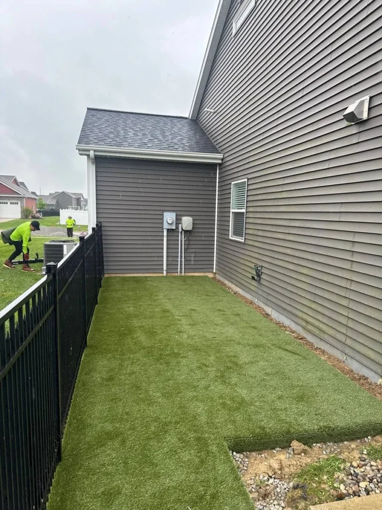 K9Grass artificial turf installed in narrow residential side yard beside gray vinyl-sided home with black aluminum fence.