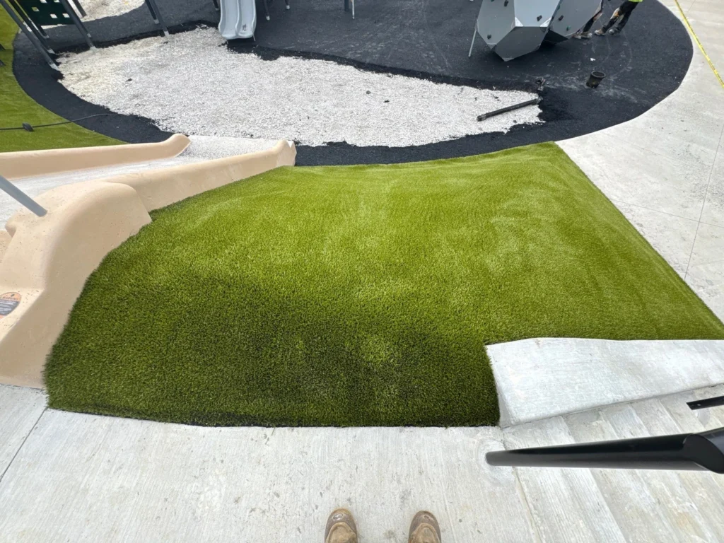 Artificial turf being installed beneath playground slide at City Lake Park, with rubber border and gravel base visible.