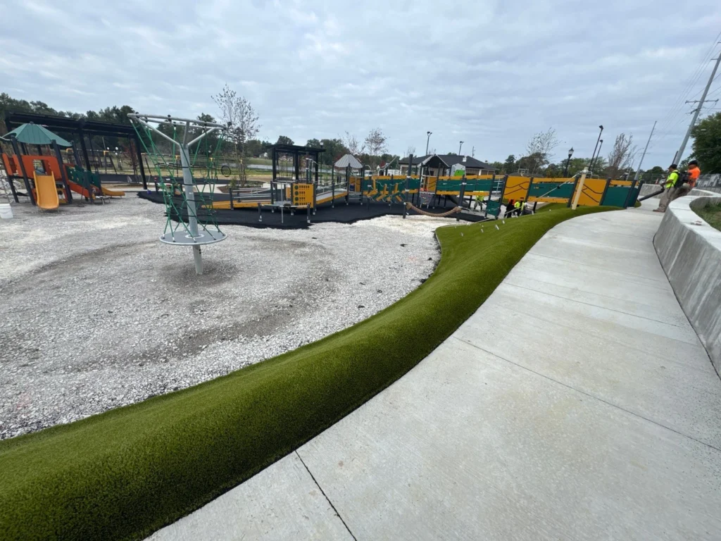 Artificial turf border installation along curved concrete path at City Lake Park playground, Booneville, with gravel and play structures beyond.