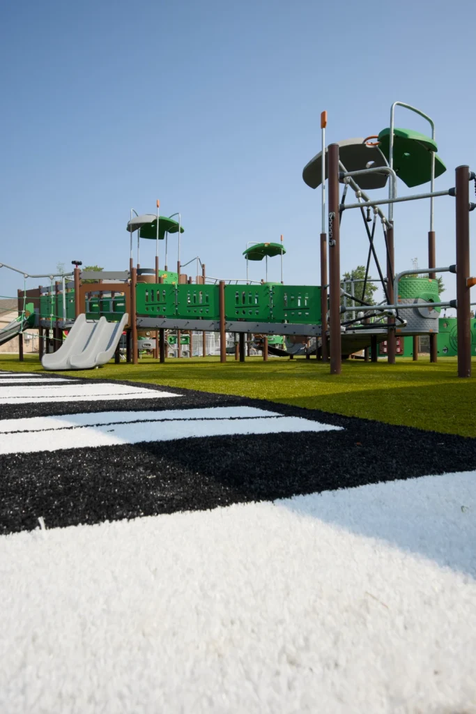Modern municipal playground with green equipment on artificial turf, bordered by black and white rubber mulch piano key pattern.