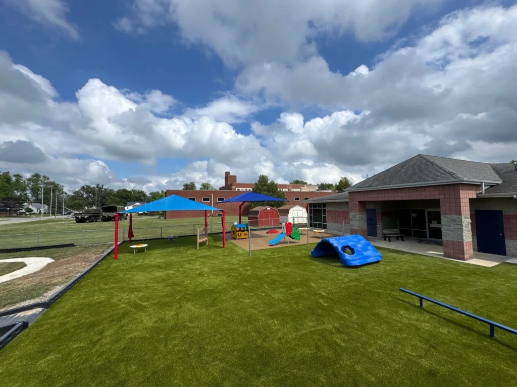 Artificial turf playground at Portland Head Start school with shade canopies, colorful play equipment, and brick building under blue sky.