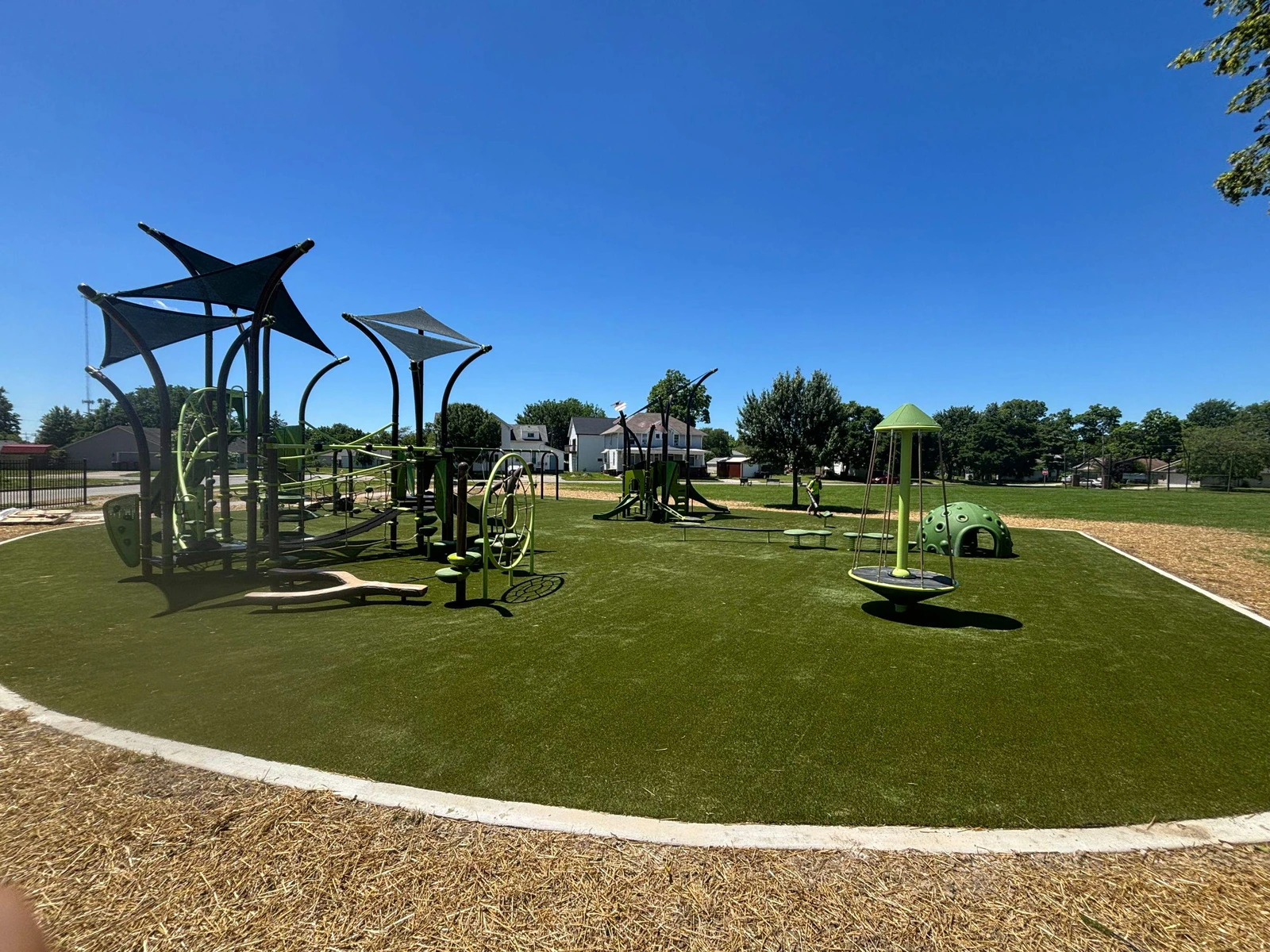 Green synthetic playground turf installed at Hanna Homestead Park with modern play structures, shade sails, and wood chip border surround.
