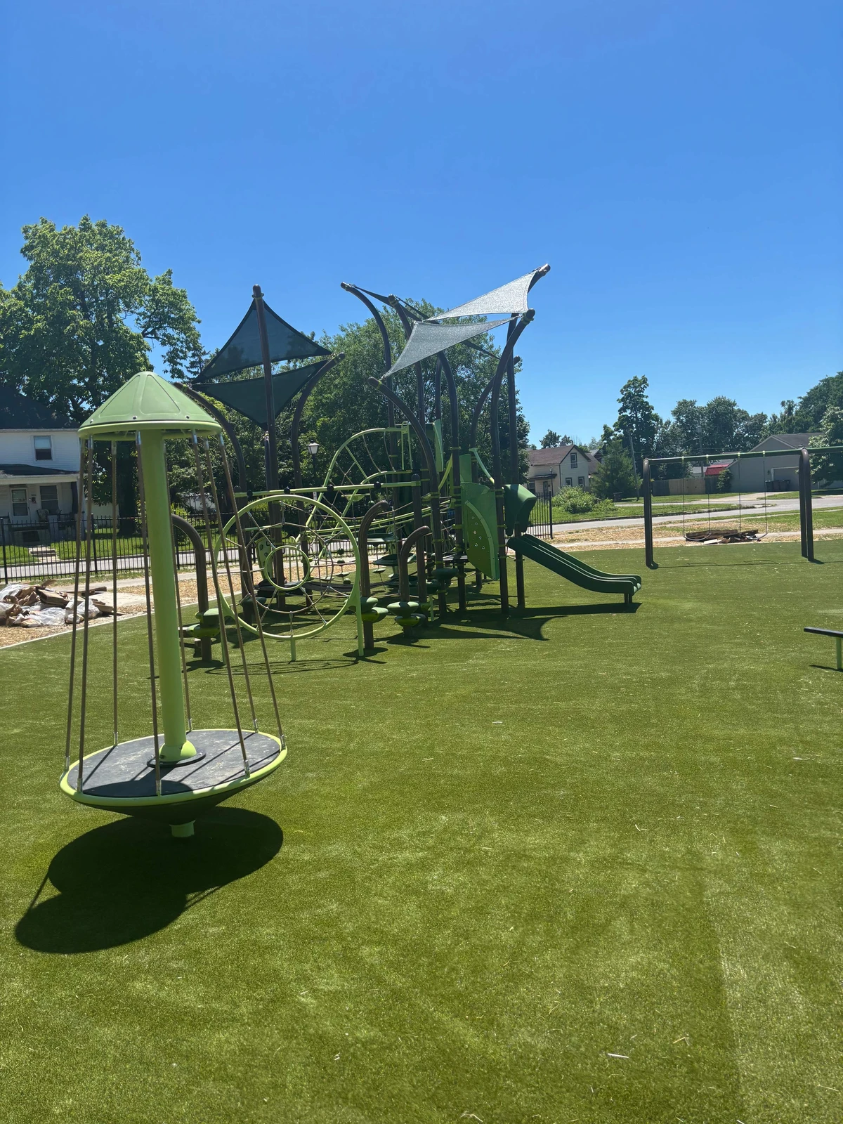 Green artificial turf surrounds a large municipal playground with climbing structures, shade sails, slide, and swings under clear blue sky.
