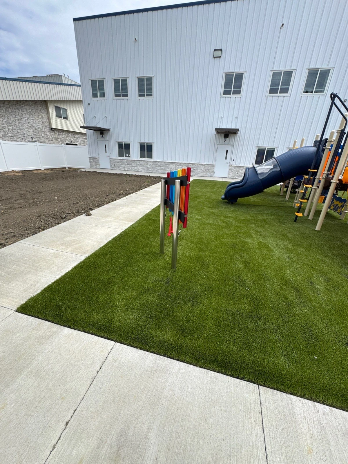 Artificial playground turf installation with colorful chimes, blue tube slide, and play structure beside a white commercial building.