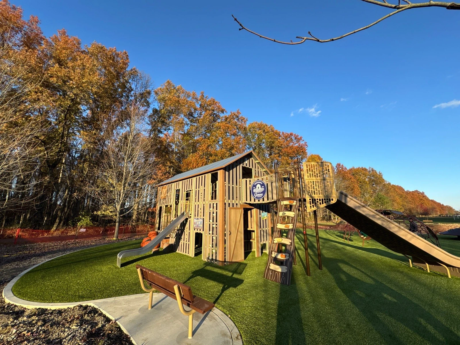 Barn-style playground with slides on synthetic turf at McNamara Park, surrounded by autumn trees under blue sky, Galena OH.