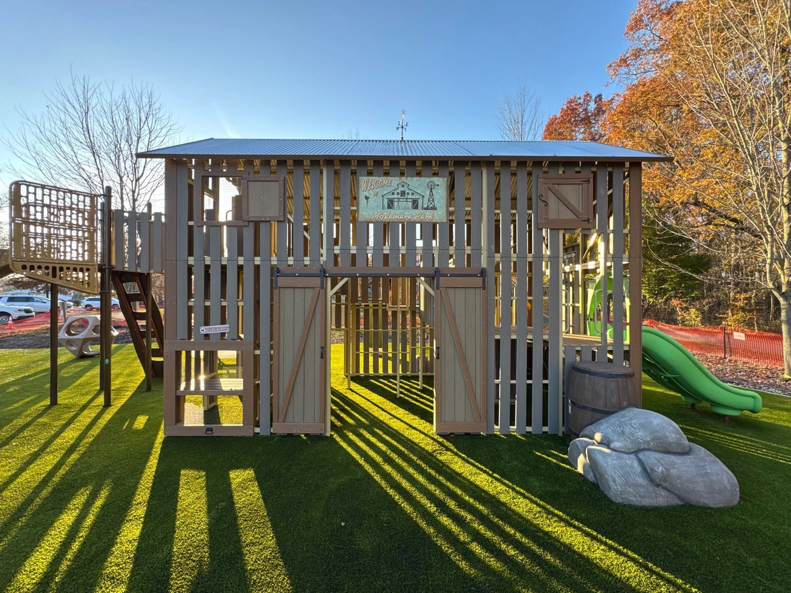 Barn-themed playground structure with artificial turf at McNamara Park, Galena OH, featuring slides, climbing walls, and a metal roof.