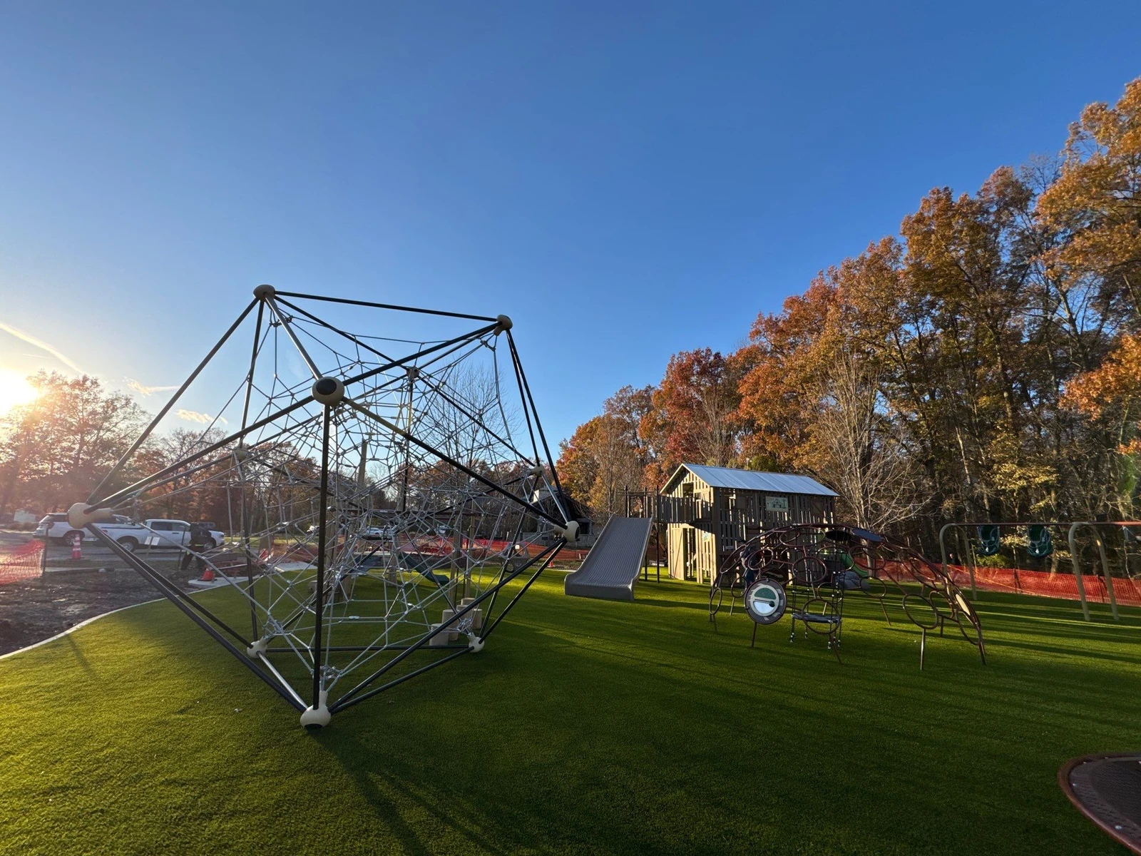Modern playground with geometric rope climber and wooden play structure on lush artificial turf, fall trees, Galena OH park.
