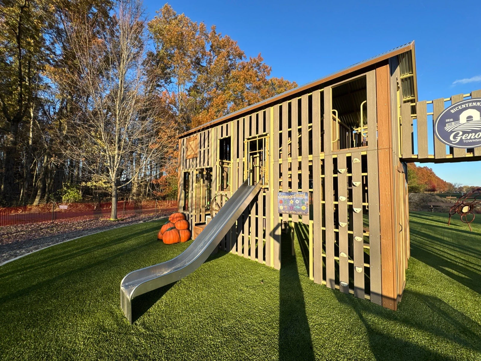 Playground Grass Ultra artificial turf beneath wooden barn-style play structure with metal slide, pumpkins, and rock wall in Galena, OH park.