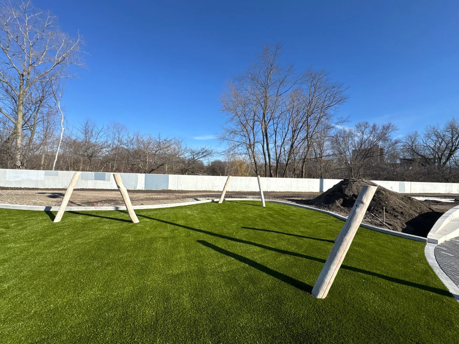 Freshly installed artificial playground turf with wooden post supports inside a concrete perimeter wall at Fort Wayne Riverfront park.