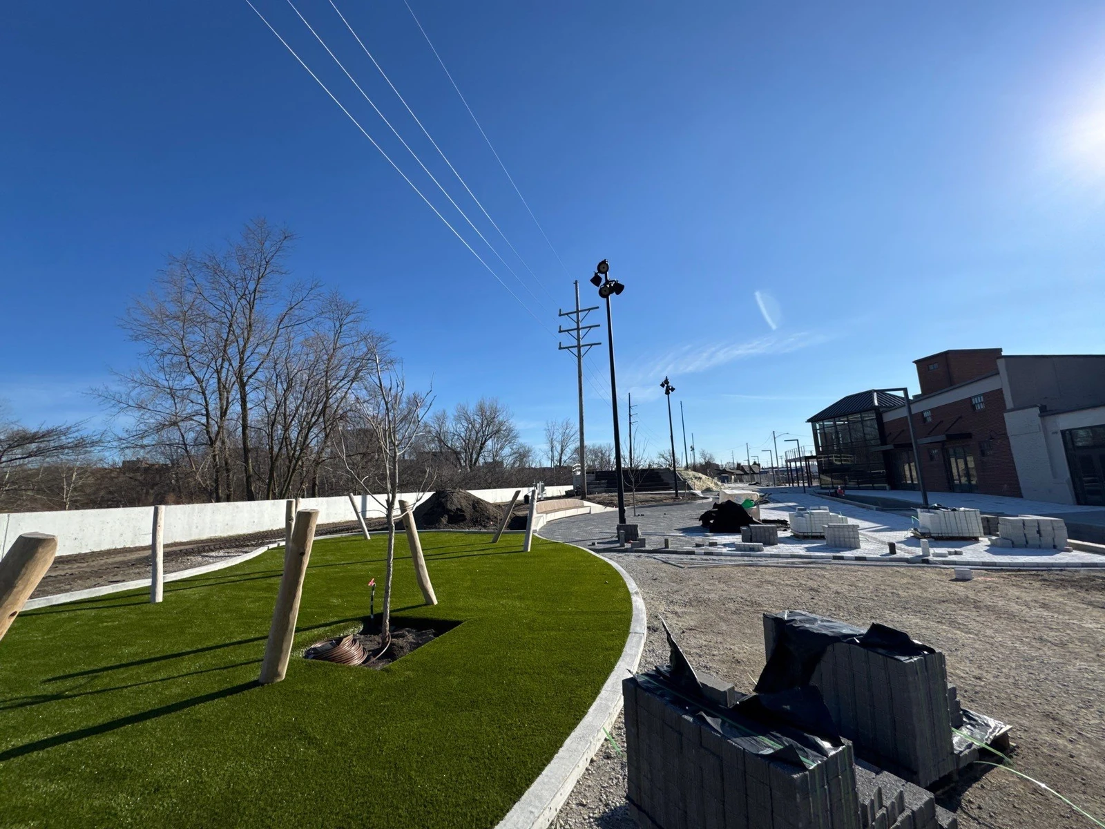ForeverLawn Playground Grass Ultra installed in curved municipal park area with timber play structures, pavers, and brick building under blue sky.