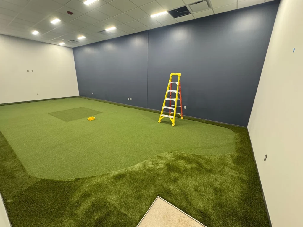 Indoor golf simulator room under construction with GolfGreens artificial putting green installed, blue accent wall, yellow ladder, Fort Wayne.