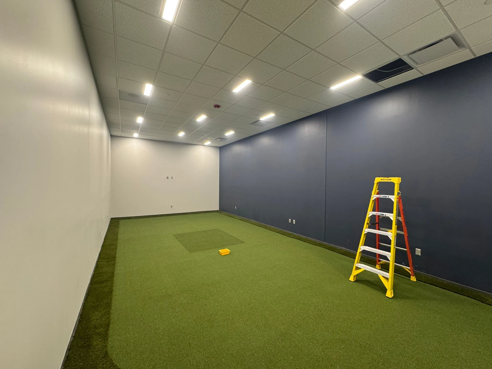 Indoor golf simulator room under construction with GolfGreens artificial turf floor, navy accent wall, drop ceiling, and yellow ladder.