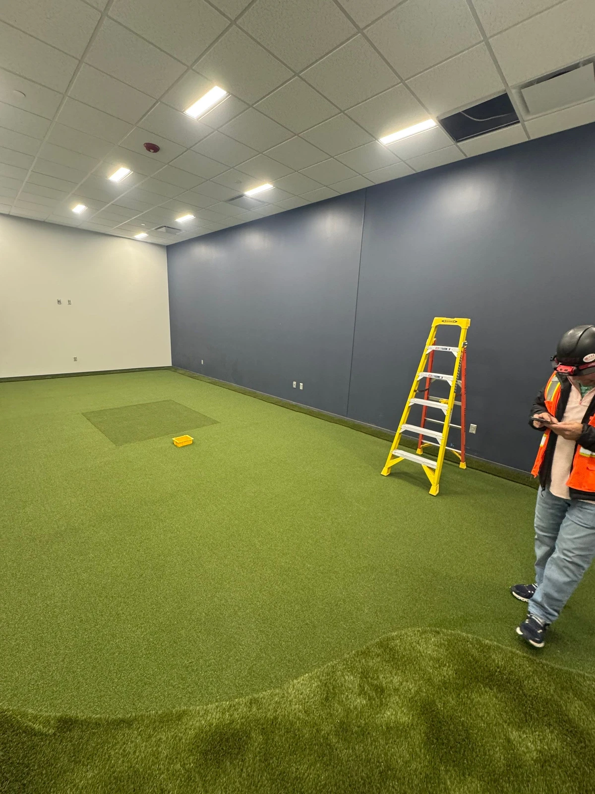 Indoor GolfGreens artificial turf installation in progress, large room with navy walls, yellow ladder, and contractor on site.