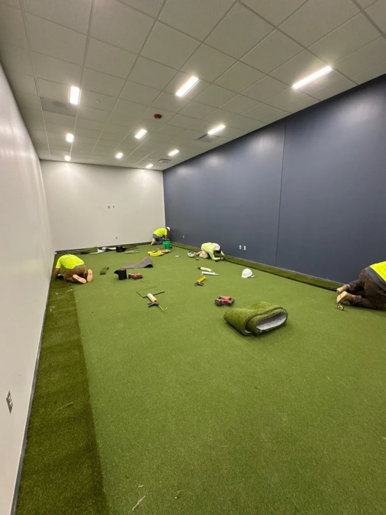 Workers in safety vests installing GolfGreens artificial turf indoors in a Fort Wayne golf simulator room with blue accent walls.