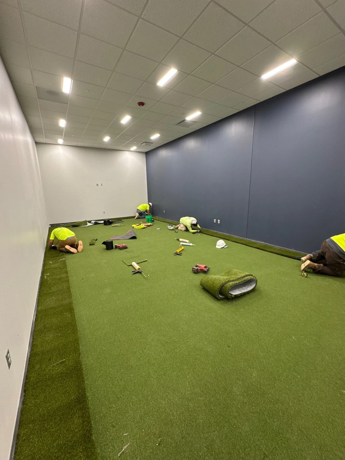 Workers in safety vests installing GolfGreens artificial turf indoors in a Fort Wayne golf simulator room with blue accent walls.