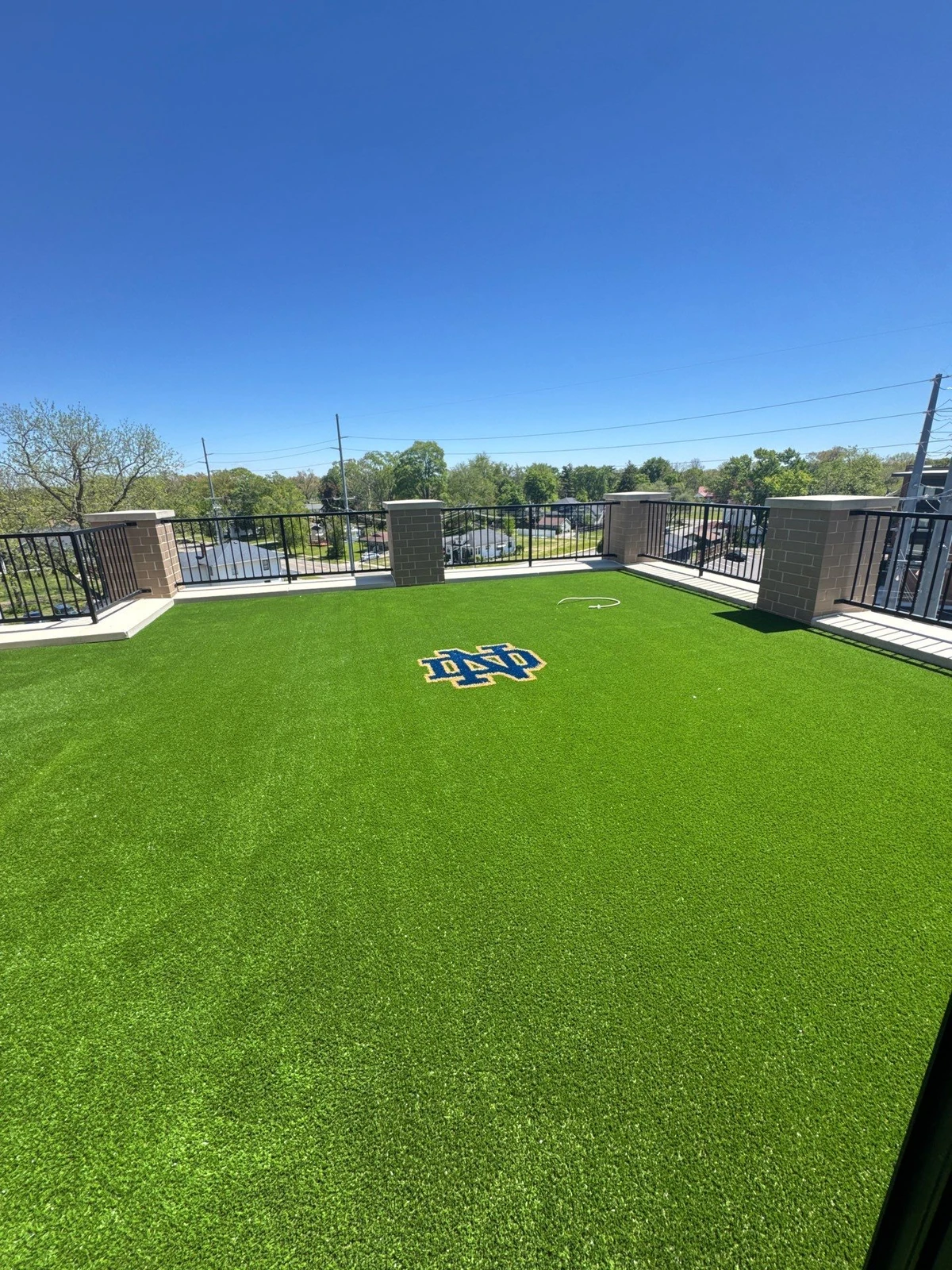 Rooftop artificial turf installation with Notre Dame ND logo inlay, 690 sq ft, black metal railing surround, clear blue sky.